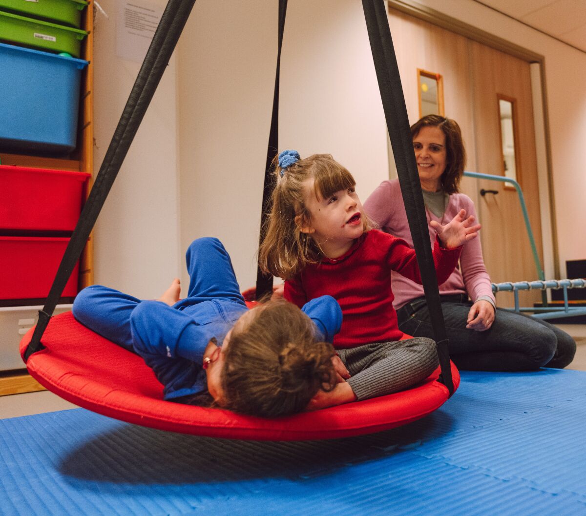 Twee kinderen spelen binnen in een schommelmand met een begeleider erbij Twee kinderen spelen binnen in een schommelmand met een begeleider erbij