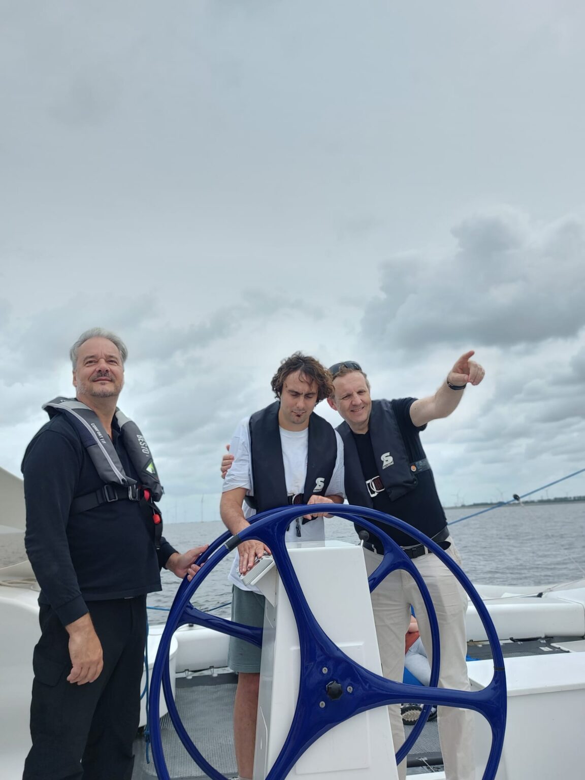 Een jongen staat met zijn ogen dicht op een zeilschip. Hij houdt het stuur met zijn handen vast. Links en rechts naast hem staan twee mannen met reddingsvesten. Een van de mannen wijst naar een plek op het water.