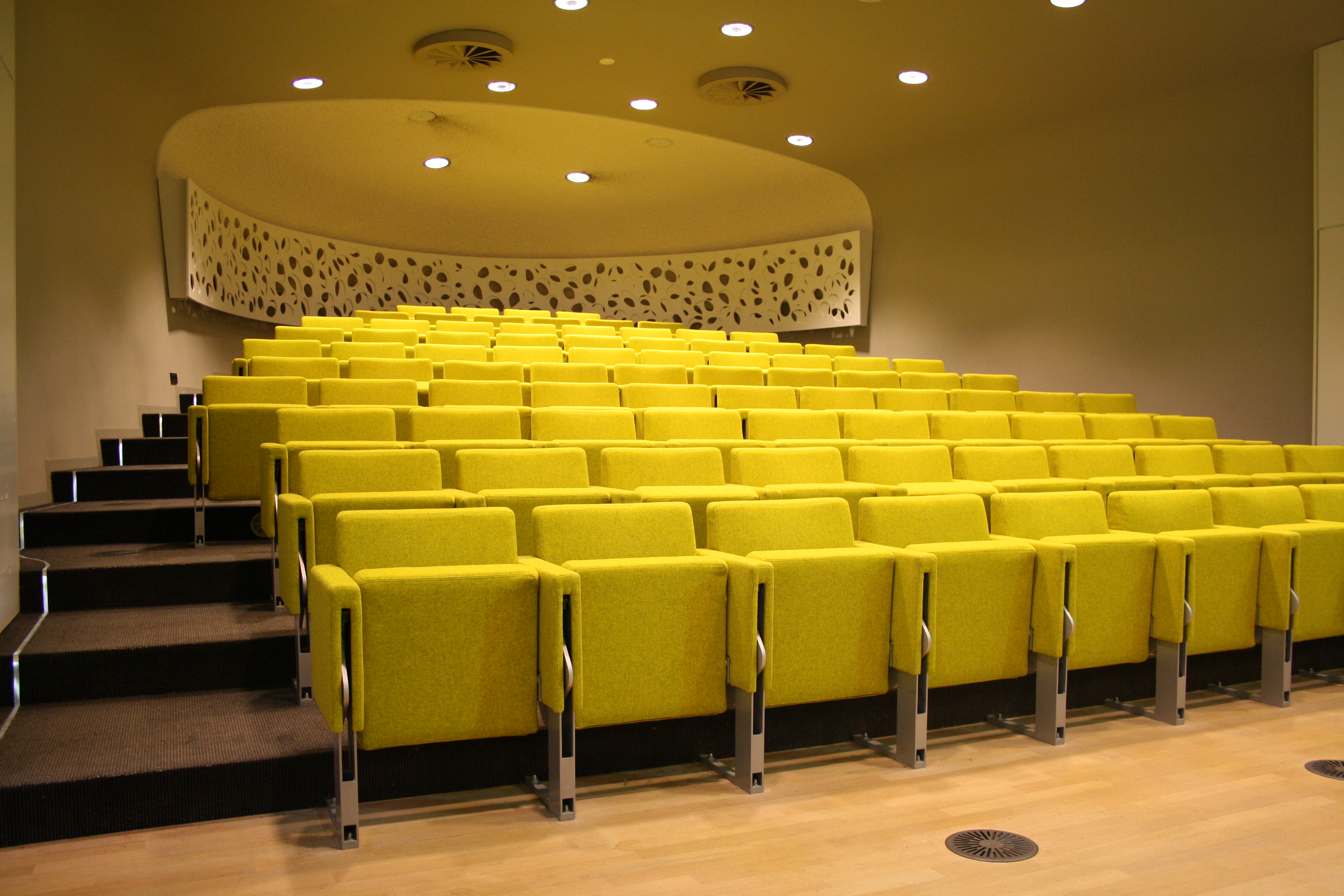 Op de foto is een congreszaal te zien, vol met gele stoelen. Op de foto is een congreszaal te zien, vol met gele stoelen.