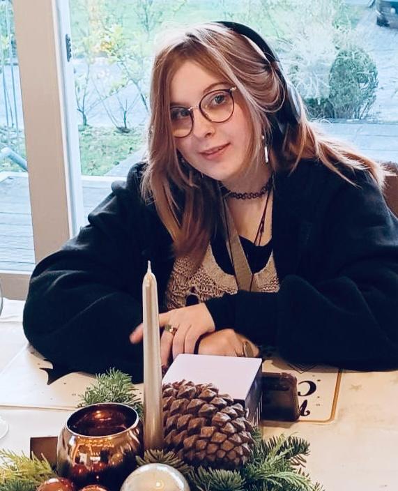 Elsie poseert glimlachend terwijl zij aan een tafel zit met een kerststukje erop, met een kaars, dennenappel en kerstgroen. Elsie draagt een koptelefoon.