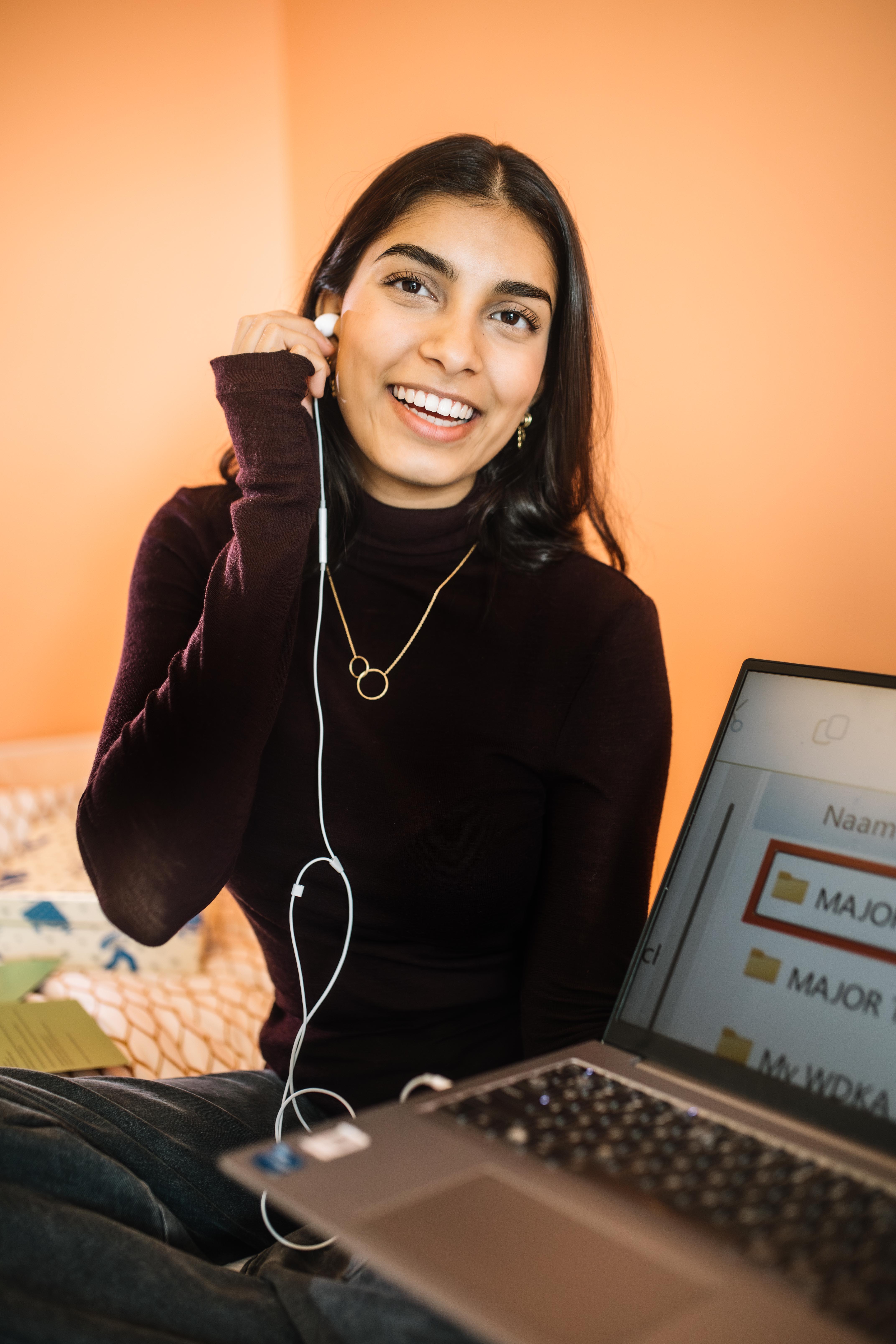 Bodhi zit op haar bed met haar laptop op schoot. Ze doet een oordopje in, en glimlacht naar de camera. Bodhi zit op haar bed met haar laptop op schoot. Ze doet een oordopje in, en glimlacht naar de camera.