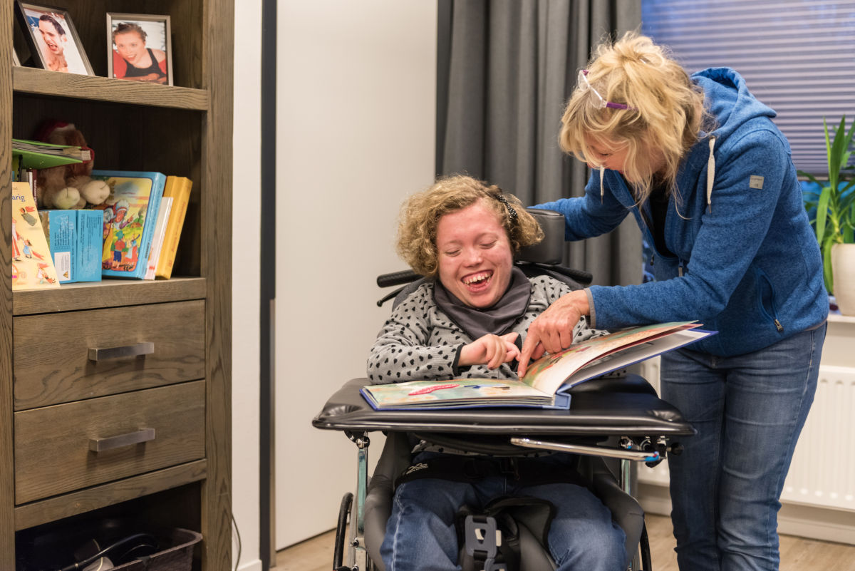 Medewerker leest een groot boek samen met een cliënt in een rolstoel, waarbij ze dingen aanwijzen in het boek Medewerker leest een groot boek samen met een cliënt in een rolstoel, waarbij ze dingen aanwijzen in het boek