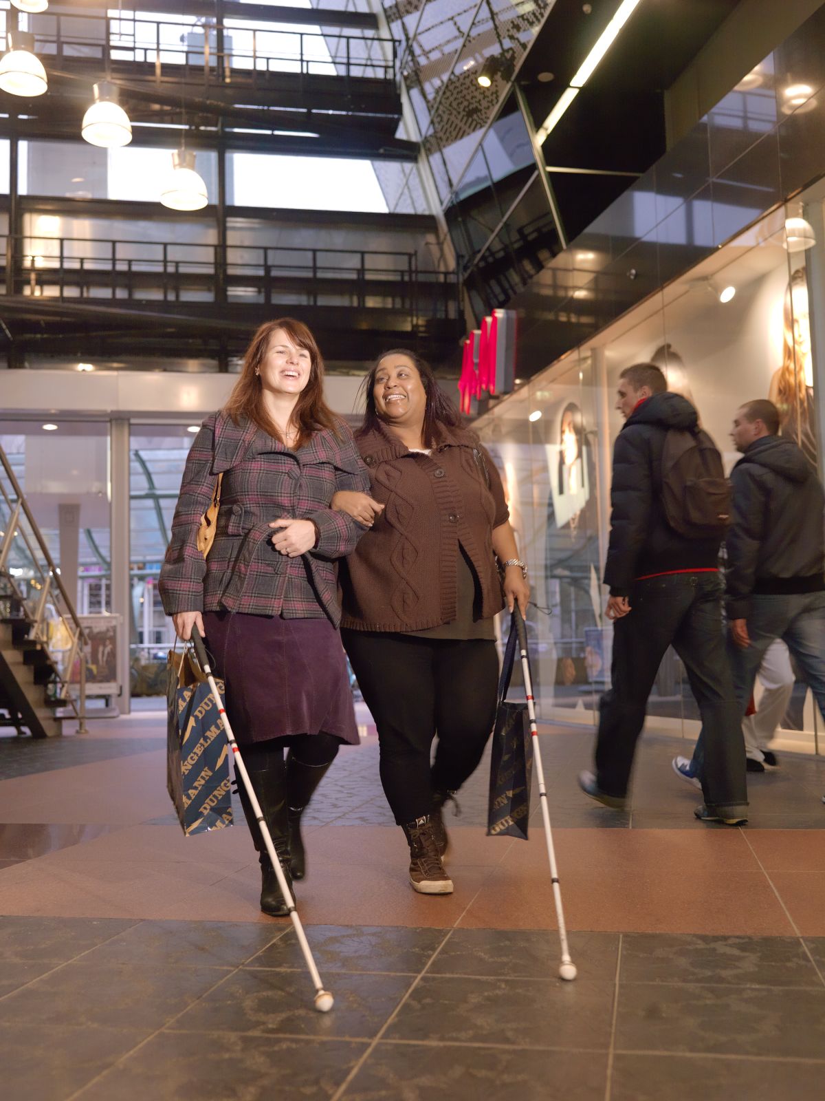 Twee vrouwen lopen door een gebouw. Ze hebben allebei een taststok vast. Twee vrouwen lopen door een gebouw. Ze hebben allebei een taststok vast.