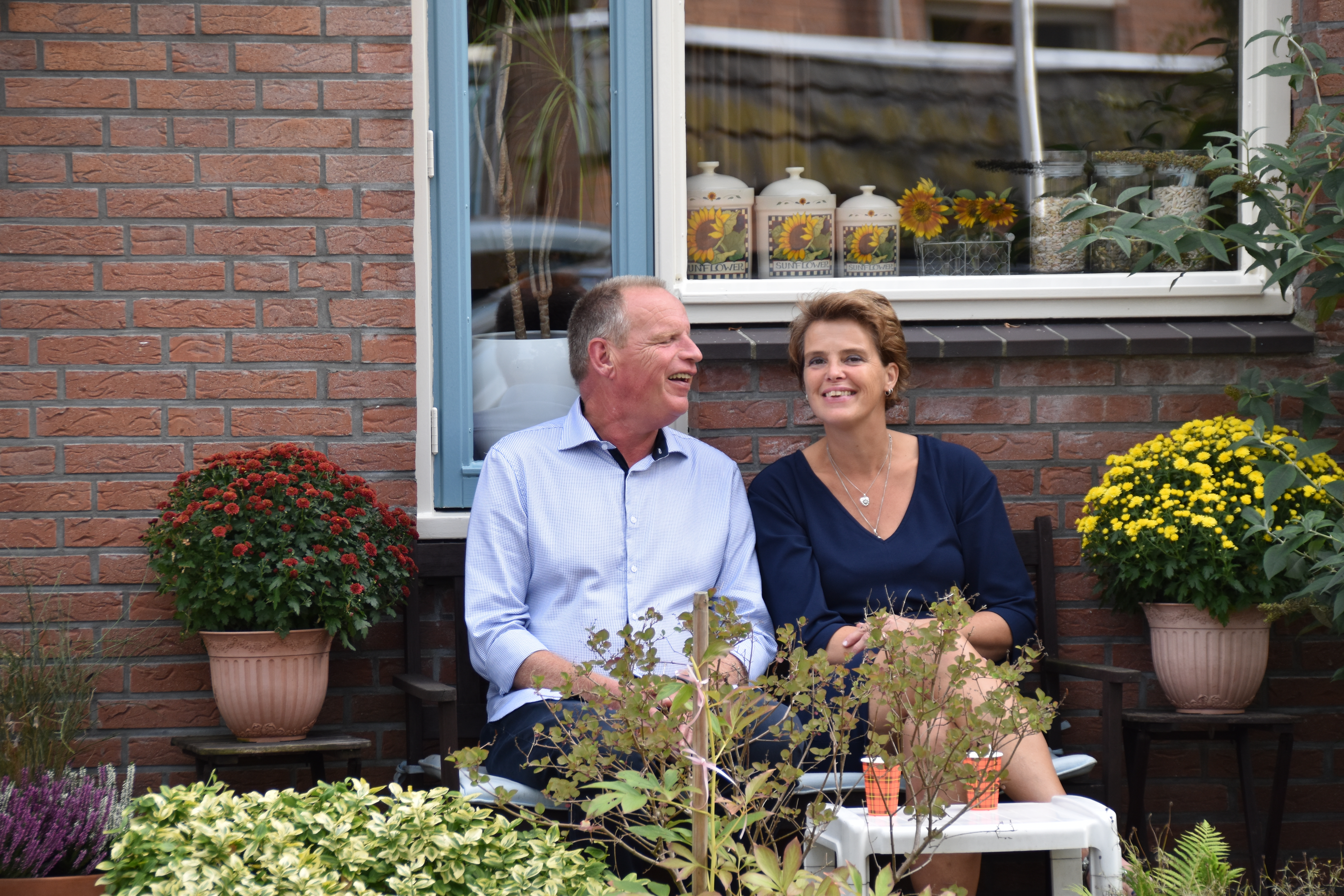 Jan zit naast Renate op een bankje in de tuin met bloemen om hen heen. Ze lachen allebei.