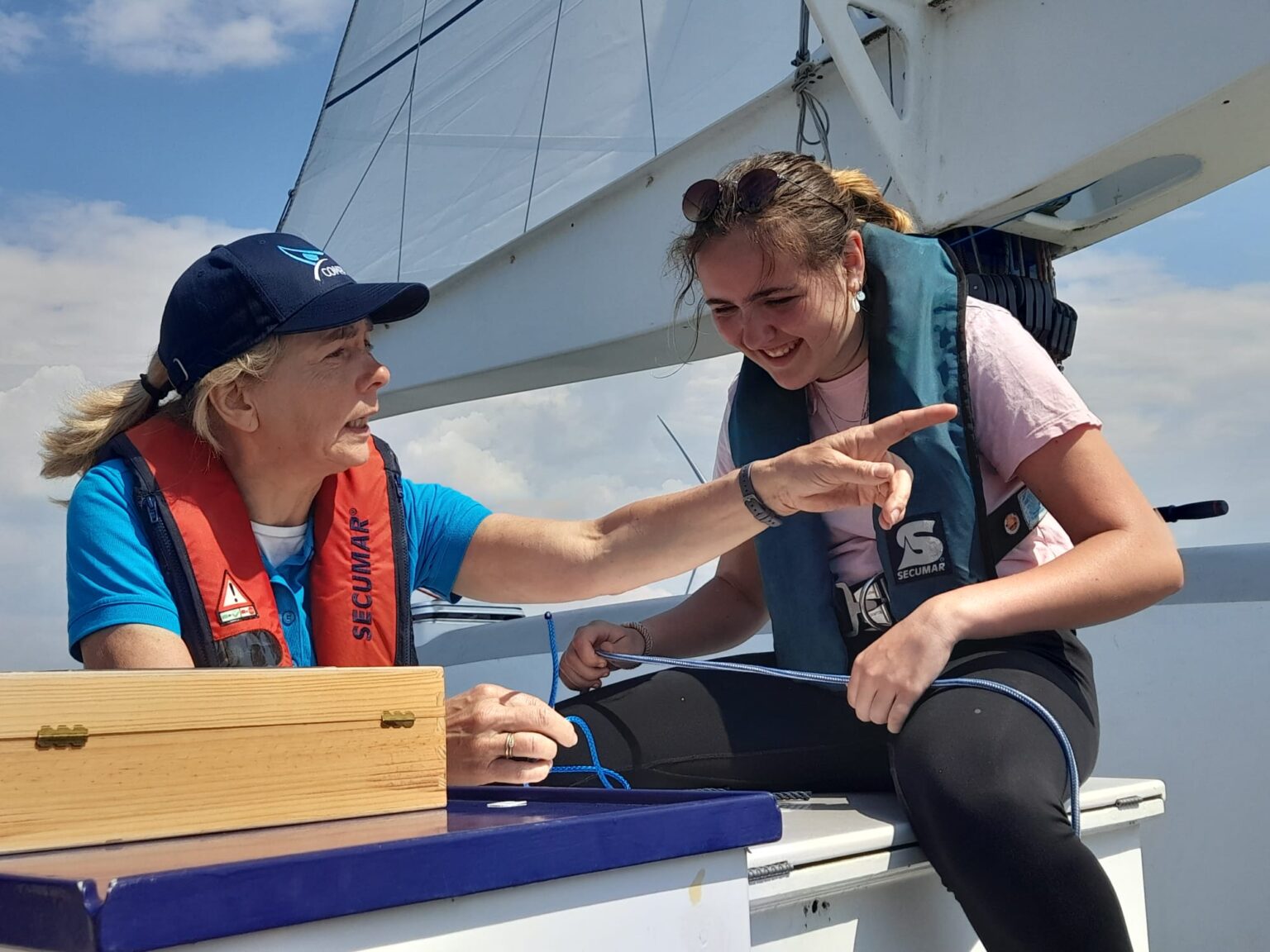 Een vrouw zit links op de zeilboot met een reddingsvest aan en pet op en wijst met haar vinger. Naast haar zit een jonge vrouw met een reddingsvest, zij heeft een touw in haar handen.