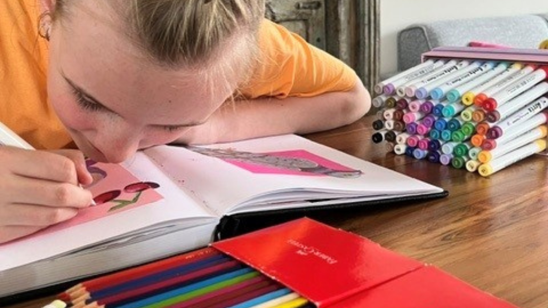 Liv zit aan een tafel en maakt een tekening in een schetsboek, omringd door stiften en kleurpotloden. Liv zit aan een tafel en maakt een tekening in een schetsboek, omringd door stiften en kleurpotloden.