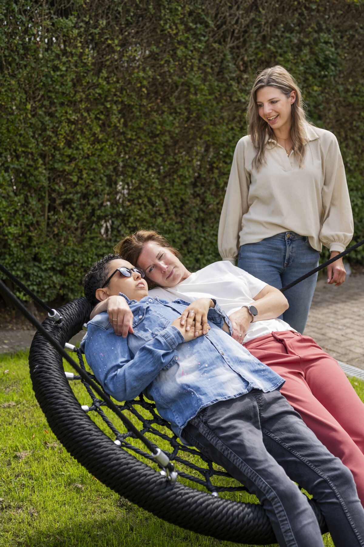 Cliënt van Visio ligt in een mandschommel en schommelt samen met een medewerker, op de achtergrond kijkt iemand glimlachend toe Cliënt van Visio ligt in een mandschommel en schommelt samen met een medewerker, op de achtergrond kijkt iemand glimlachend toe