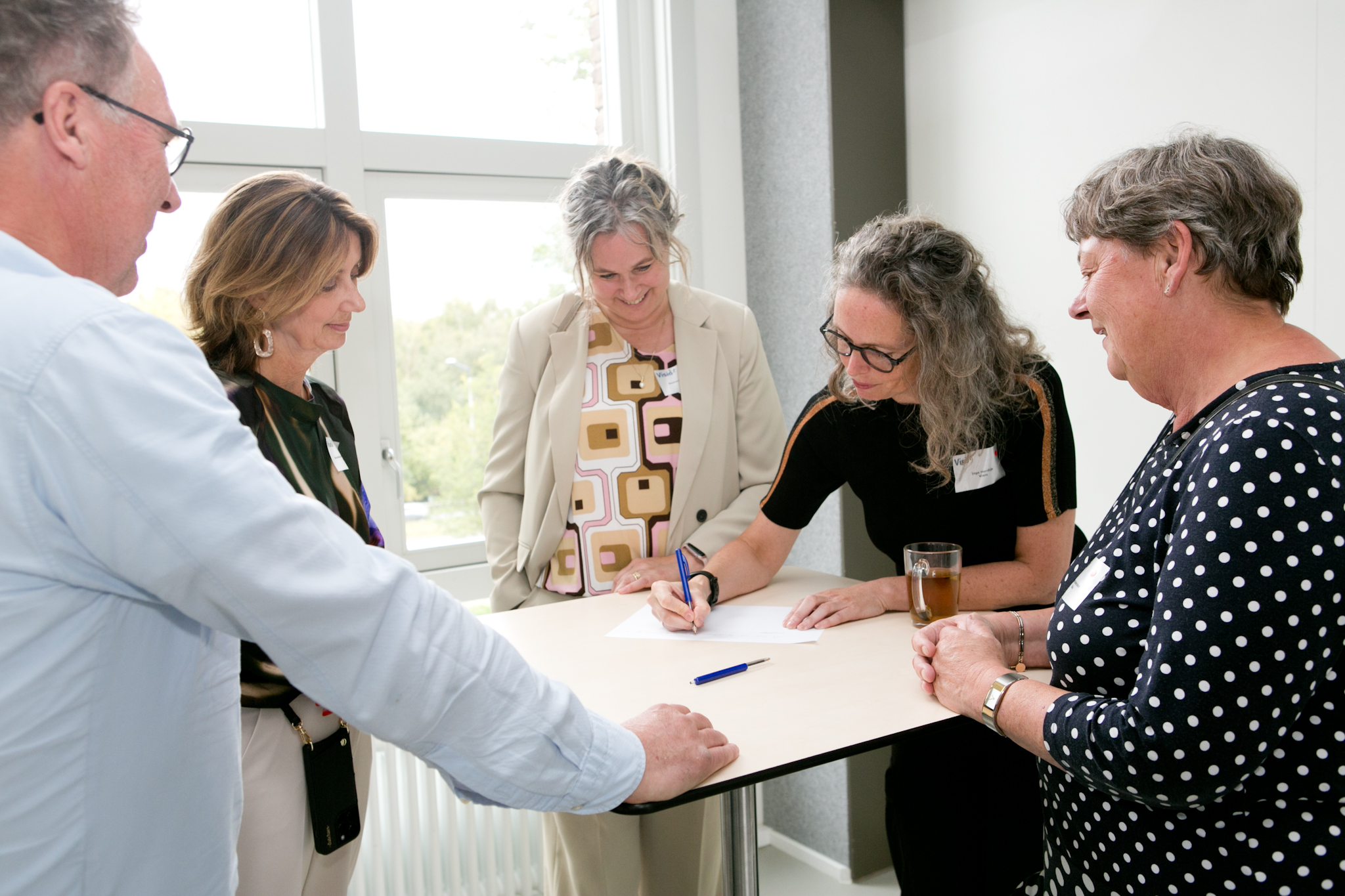 Vijf mensen staan rond een tafel en schrijven op een papier