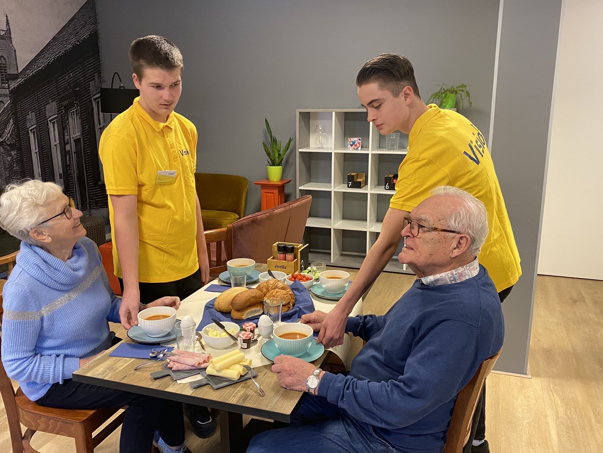 Twee leerlingen bedienen twee oudere klanten die lunchen bij huiskamercafé Visit