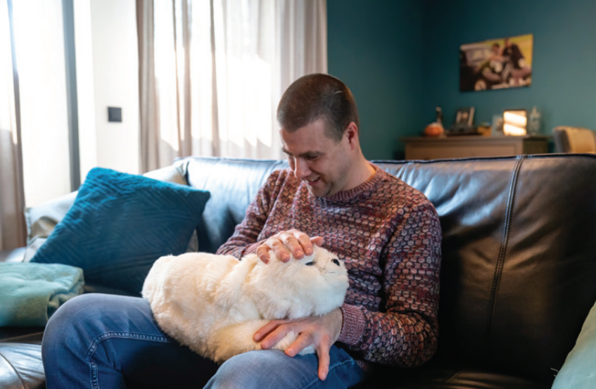 Cliënt zit op de bank met Paro de zeehond op zijn schoot. Cliënt zit op de bank met Paro de zeehond op zijn schoot.