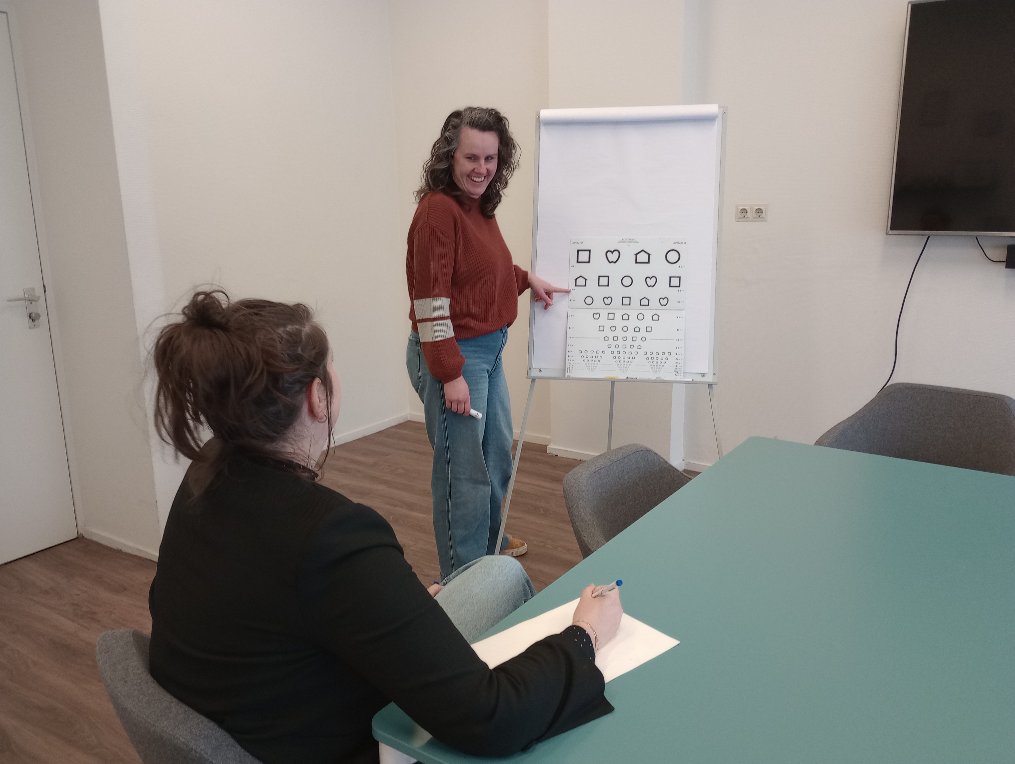 Medewerker van Visio staat voor een bord en wijst naar de symbolen die daar op staan. Ze legt iets uit aan een andere persoon, die naar het bord kijkt. Medewerker van Visio staat voor een bord en wijst naar de symbolen die daar op staan. Ze legt iets uit aan een andere persoon, die naar het bord kijkt.