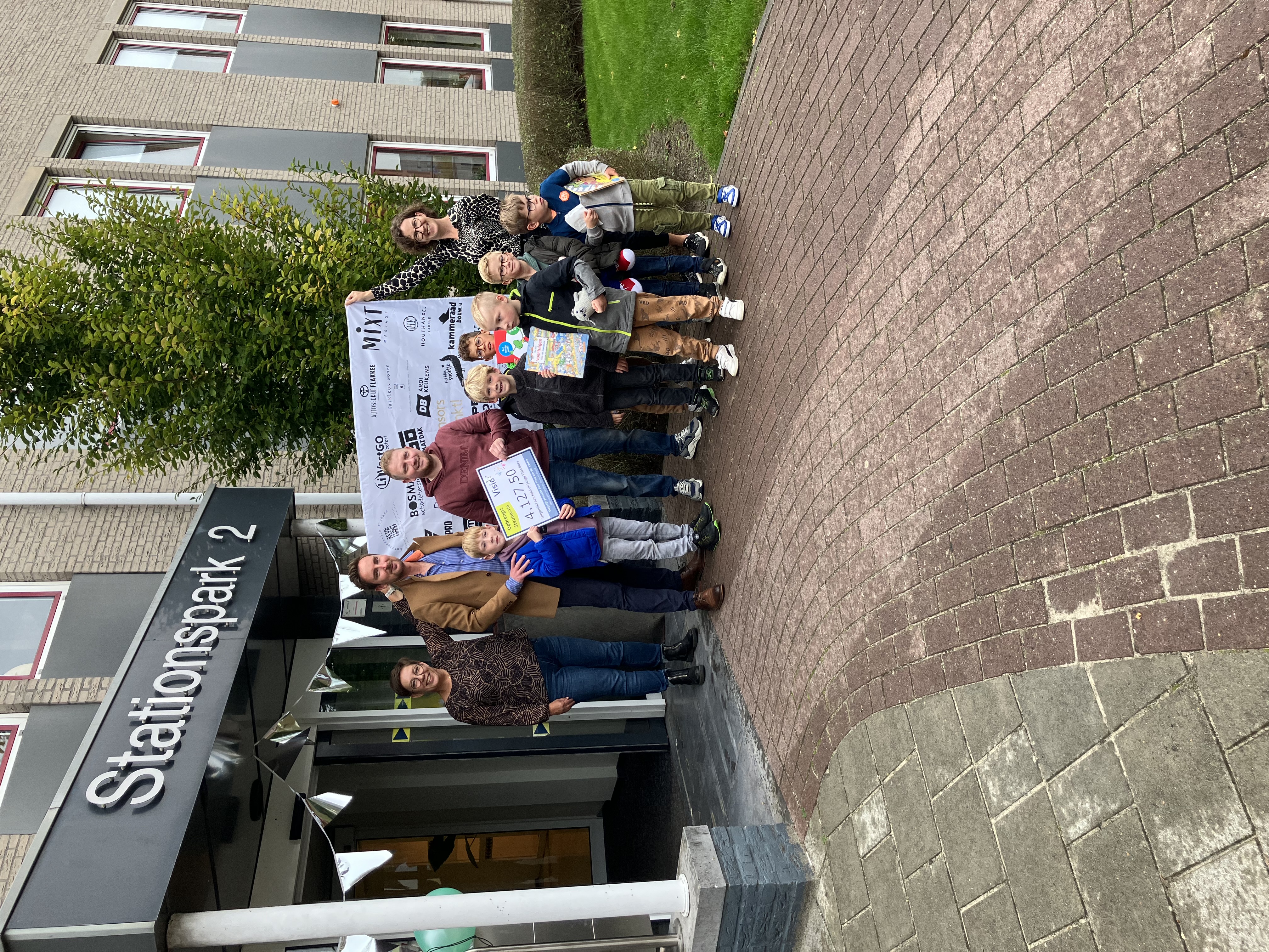 Vier volwassenen en zes kinderen poseren voor een spandoek. Theo staat ook op de foto en houdt een cheque in zijn hand vast.