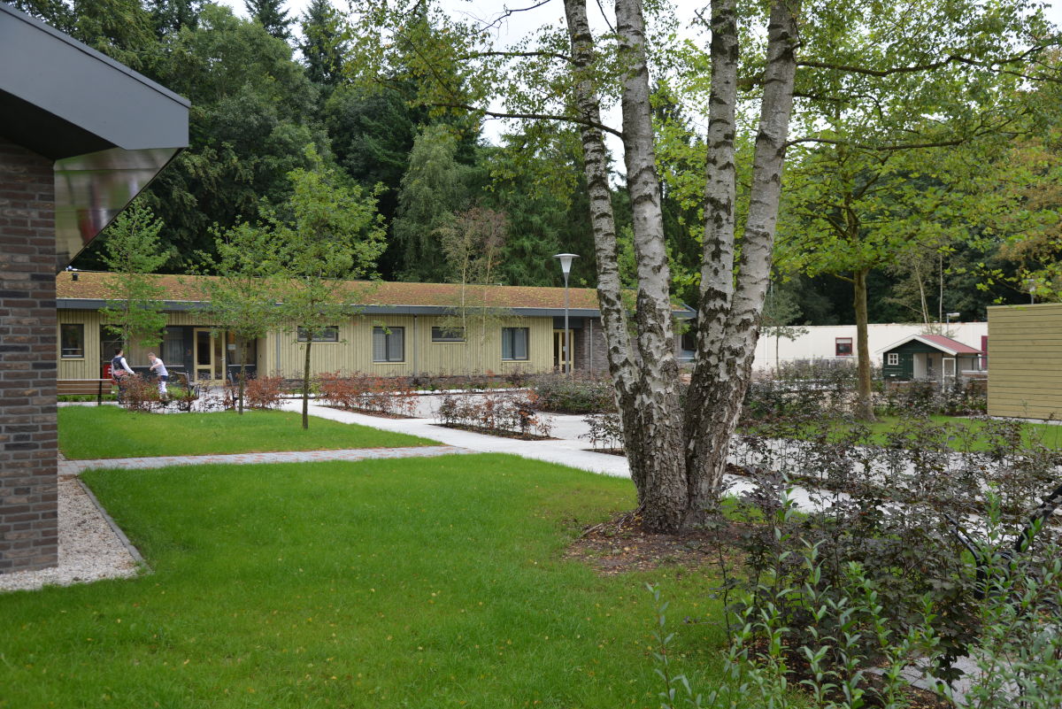 Sfeerimpressie van het groene terrein van De Blauwe Kamer met op de achtergrond de woningen en op de voorgrond veel bomen en gras Sfeerimpressie van het groene terrein van De Blauwe Kamer met op de achtergrond de woningen en op de voorgrond veel bomen en gras