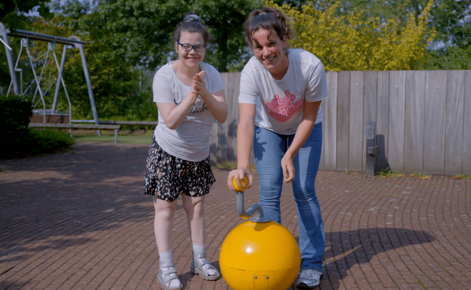 Cliënt met visuele en verstandelijke beperking en begeleider poseren bij een speeltoestel Cliënt met visuele en verstandelijke beperking en begeleider poseren bij een speeltoestel