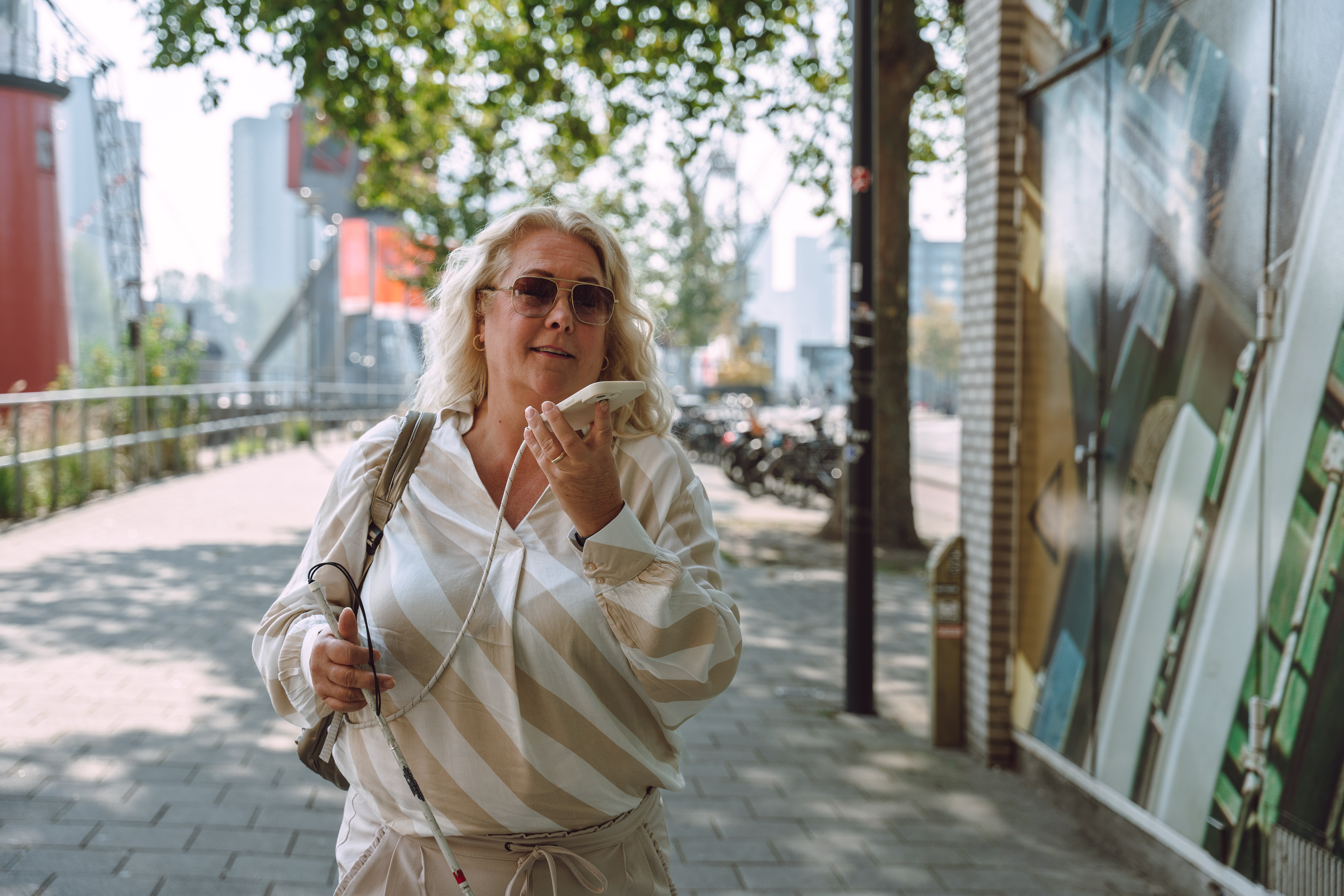 Vrouw loopt in stad, heeft een herkenningsstok in haar hand en een mobiel in haar andere hand die ze bij haar gezicht houdt. Ze draagt een bril met donkere glazen.