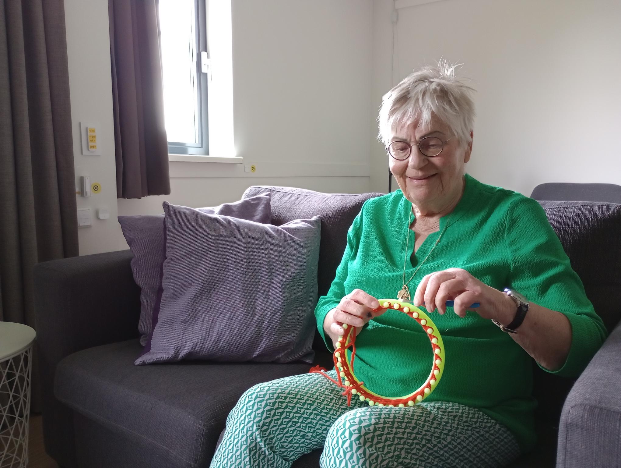 Foto van Sannie die met een breiring creatief aan de slag is, zij draagt een broek met een groen-wit patroon, een groene blouse met mouwen en een bril. Foto van Sannie die met een breiring creatief aan de slag is, zij draagt een broek met een groen-wit patroon, een groene blouse met mouwen en een bril.