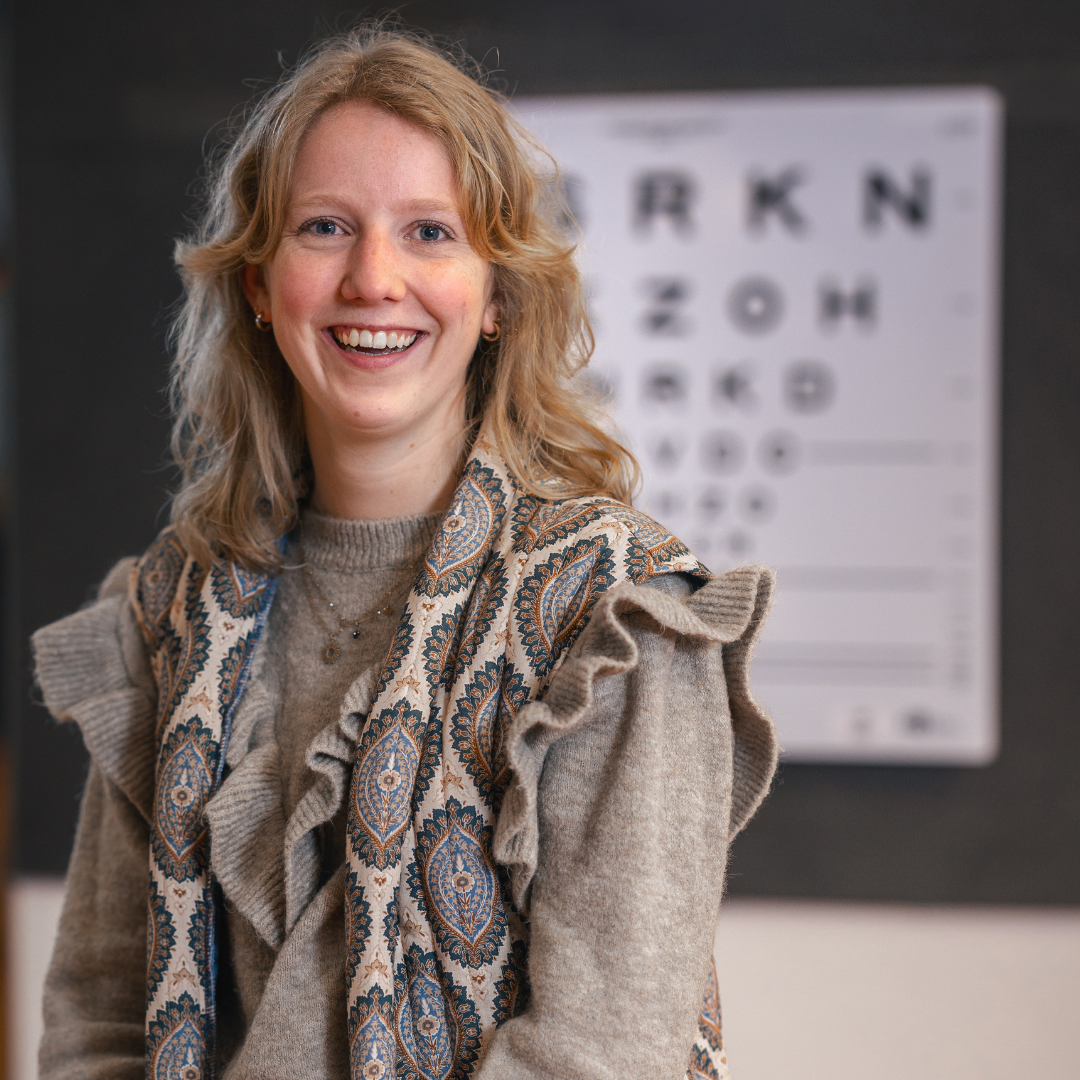 Veerle heeft blond haar tot haar schouders en kijkt lachend in de camera. Op de achtergrond zie je een oogtest hangen.