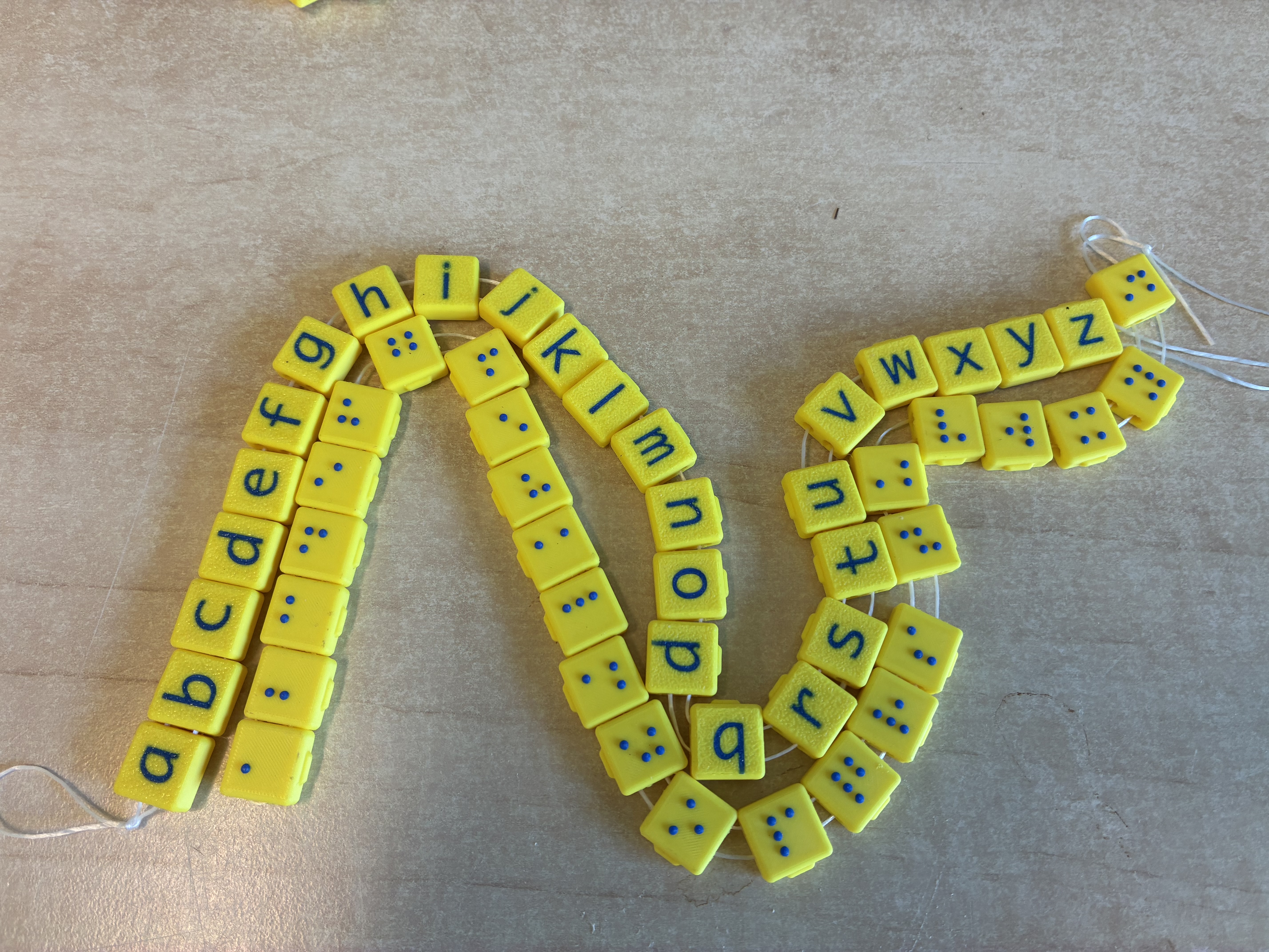 Twee koorden met gele, 3D-geprinte blokjes: één met letters van het alfabet en één met de bijbehorende brailletekens, als tastbaar leermiddel voor kinderen met een visuele beperking. Twee koorden met gele, 3D-geprinte blokjes: één met letters van het alfabet en één met de bijbehorende brailletekens, als tastbaar leermiddel voor kinderen met een visuele beperking.