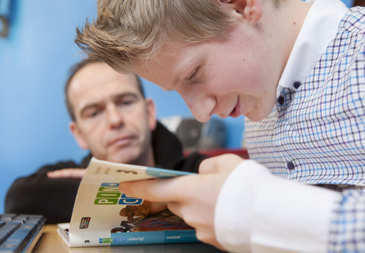 Leerling kijkt in een boek terwijl een docent meekijkt. Leerling kijkt in een boek terwijl een docent meekijkt.