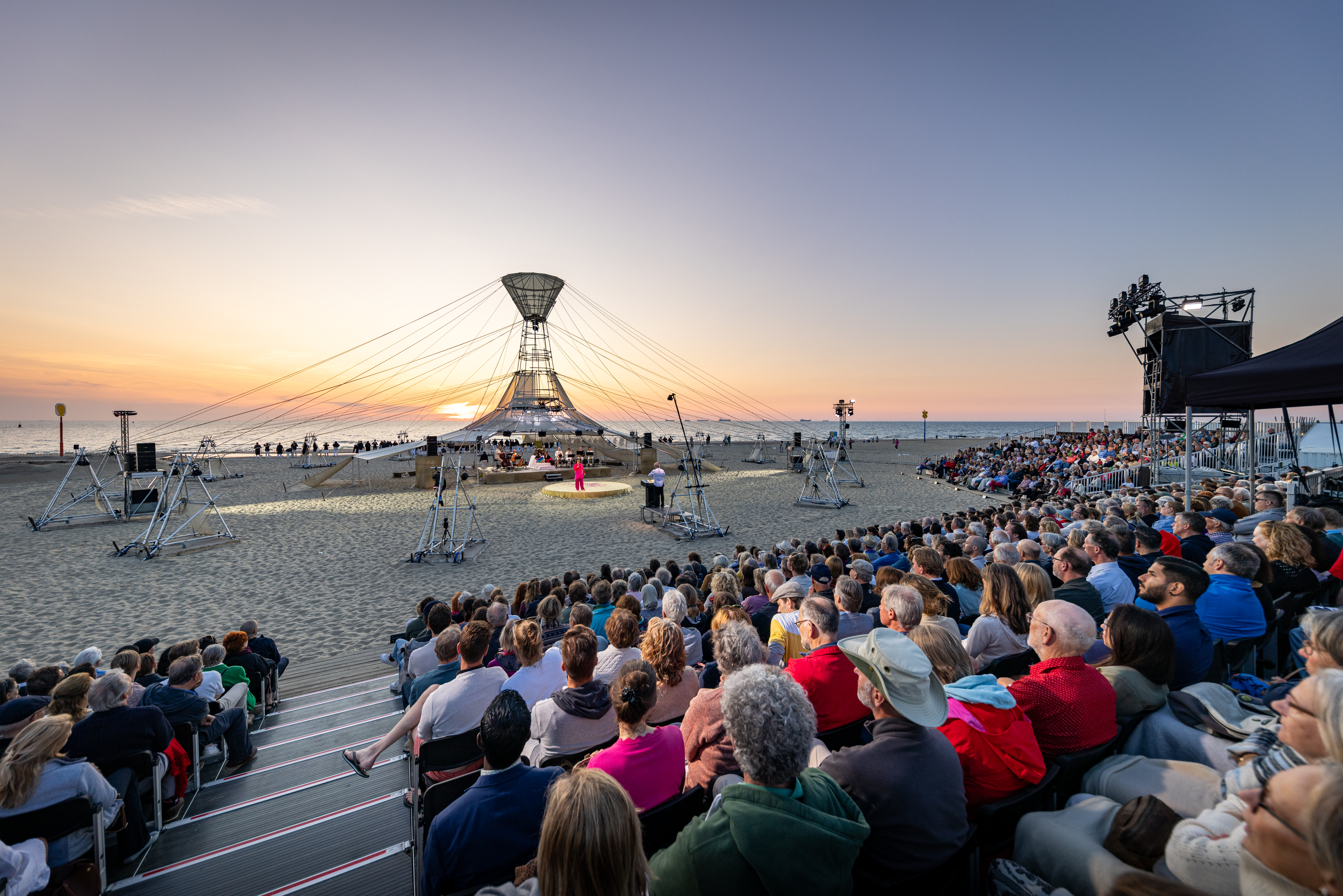 Een podium op het strand met volle tribunes eromheen en een ondergaande zon op de achtergrond