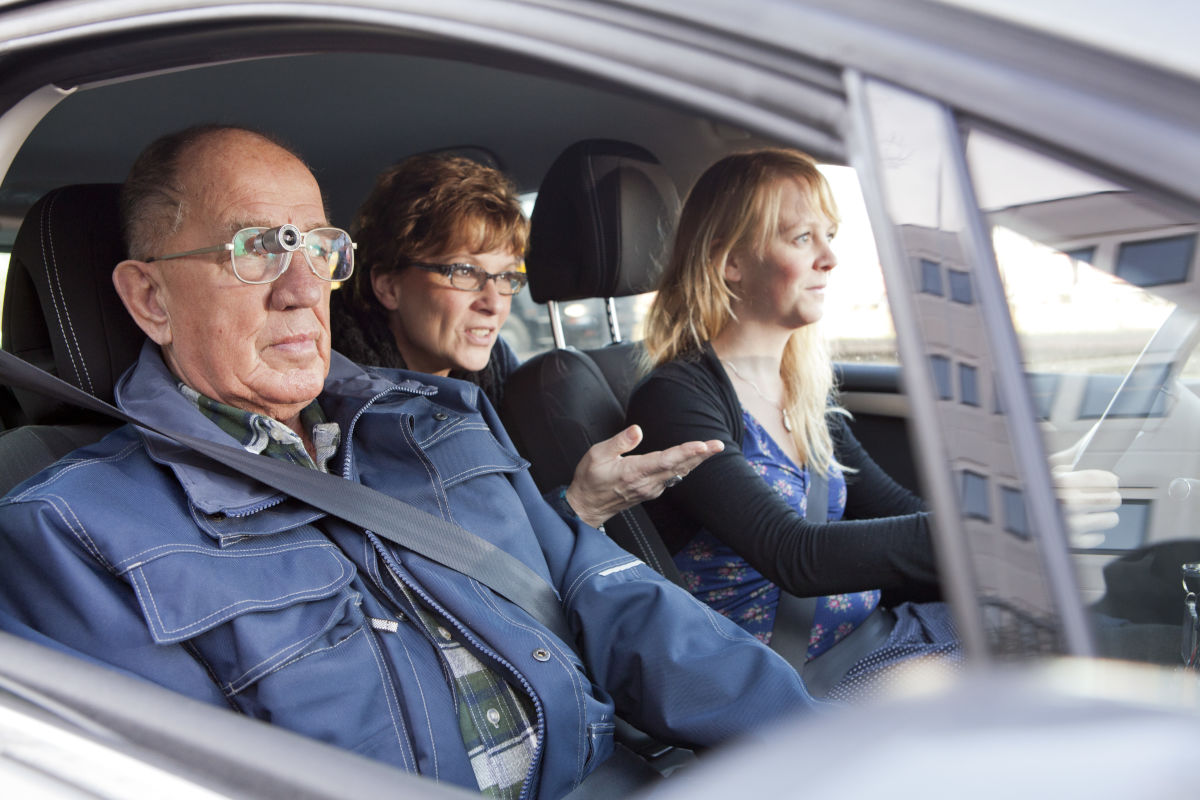 Drie personen zitten in een auto, de bijrijder draagt een bioptisch telescoopsysteem Drie personen zitten in een auto, de bijrijder draagt een bioptisch telescoopsysteem