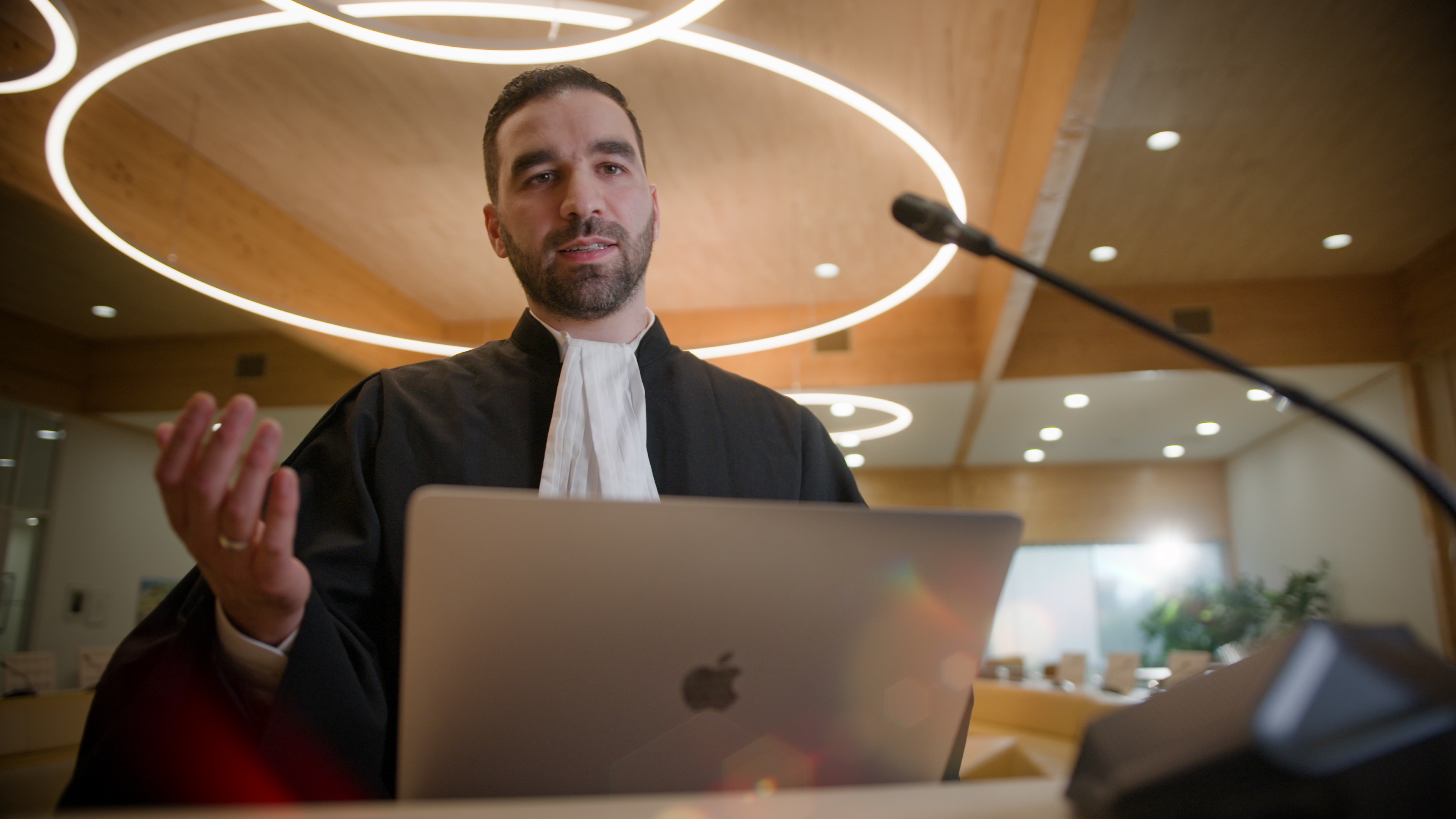 Faruk draagt zijn werkkleding (advocaat) en staat achter een microfoon. Zijn laptop staat voor hem. Hij is aan het praten. Faruk draagt zijn werkkleding (advocaat) en staat achter een microfoon. Zijn laptop staat voor hem. Hij is aan het praten.