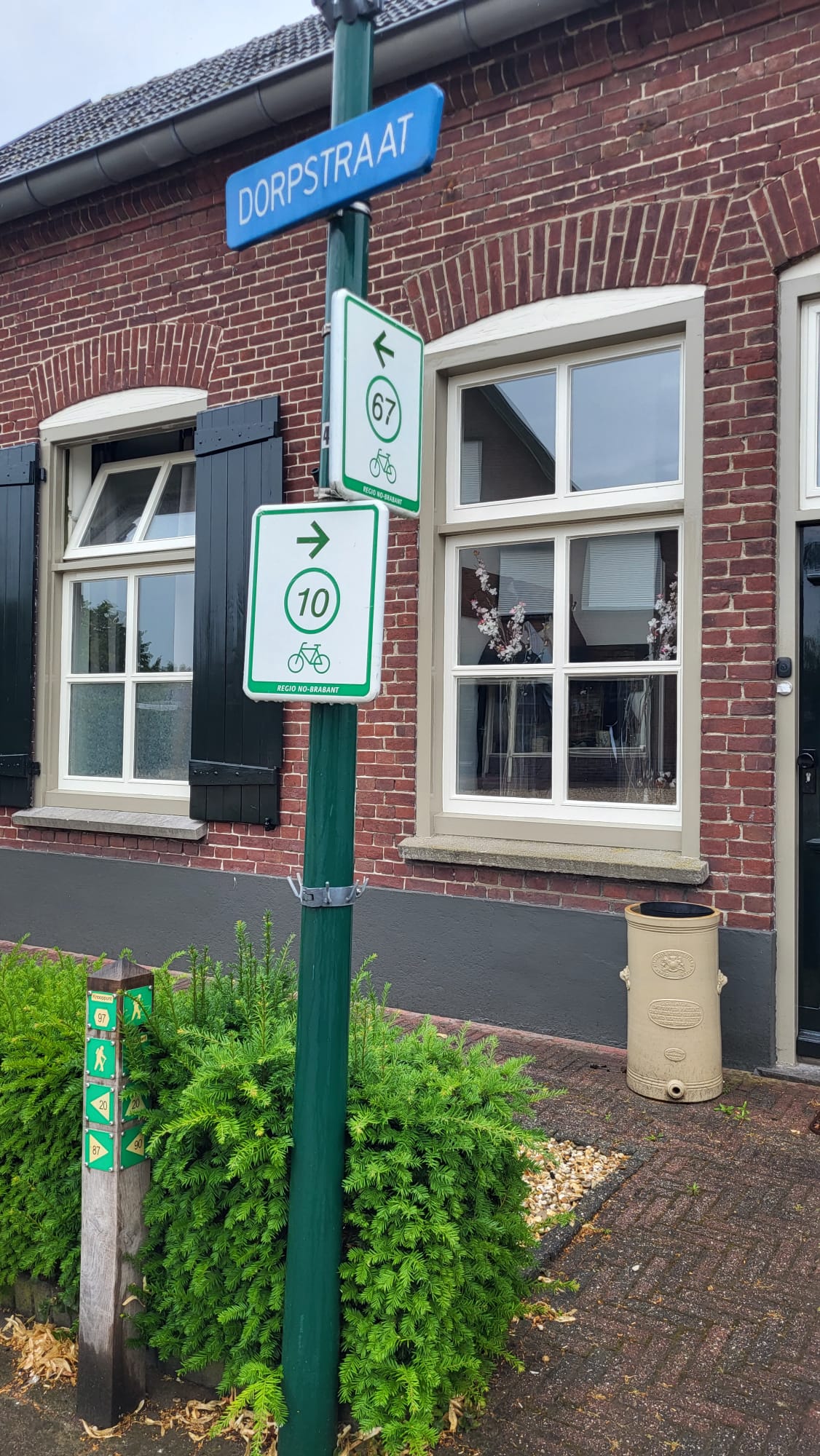 Een gebouw is te zien op de achtergrond. Op de voorgrond is een lantaarnpaal met een blauw straatnaambordje met "dorpstraat" erop. Op de paal zijn ook twee groen/witte bordjes van fietsroutes te zien. De bovenste wijst naar links en heeft het nummer 67, de onderste wijst naar rechts en heeft het nummer 10. Een gebouw is te zien op de achtergrond. Op de voorgrond is een lantaarnpaal met een blauw straatnaambordje met "dorpstraat" erop. Op de paal zijn ook twee groen/witte bordjes van fietsroutes te zien. De bovenste wijst naar links en heeft het nummer 67, de onderste wijst naar rechts en heeft het nummer 10.