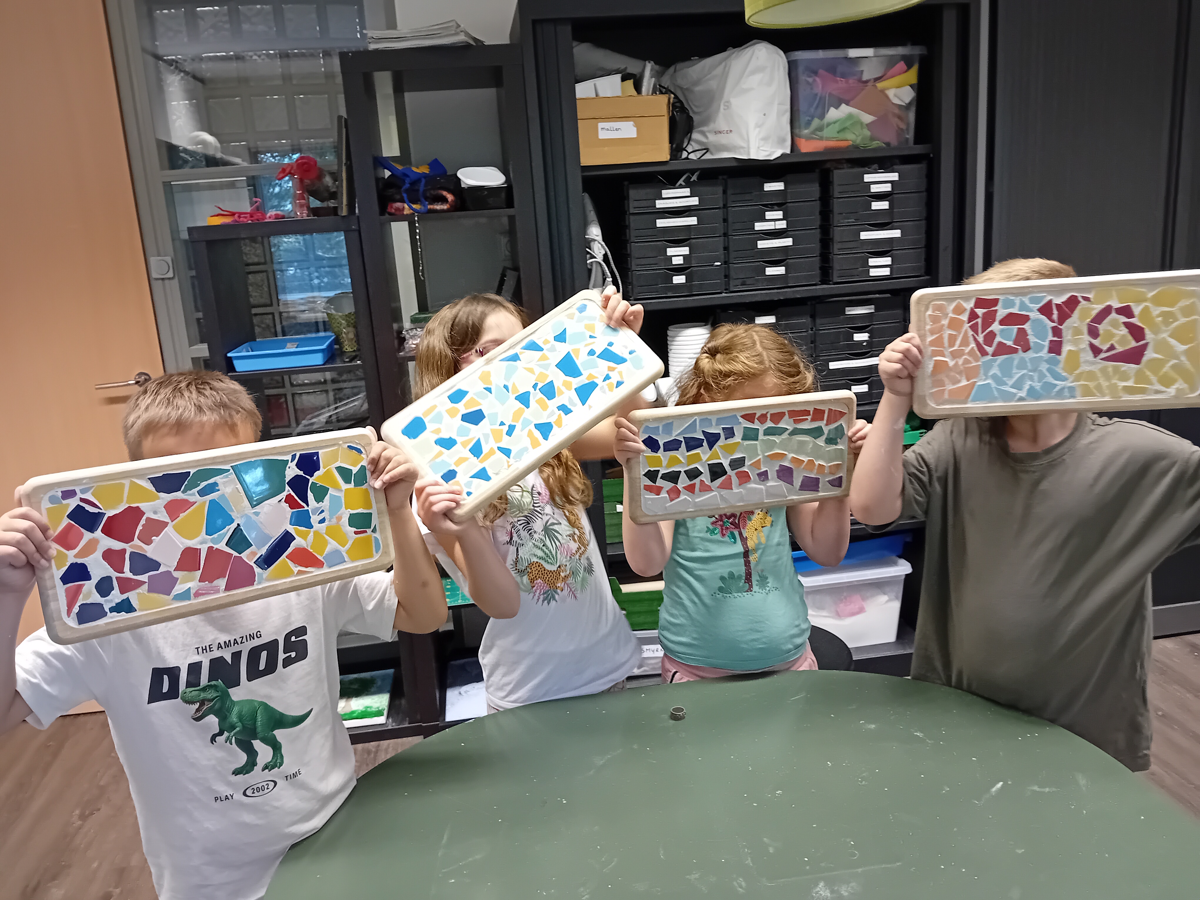 Vier kinderen houden elk een rechthoekig plateau omhoog, versierd met kleurrijke stukjes keramiek in mozaïekpatronen, gemaakt tijdens een knutselactiviteit bij Visio met kasten en materialen op de achtergrond. Vier kinderen houden elk een rechthoekig plateau omhoog, versierd met kleurrijke stukjes keramiek in mozaïekpatronen, gemaakt tijdens een knutselactiviteit bij Visio met kasten en materialen op de achtergrond.