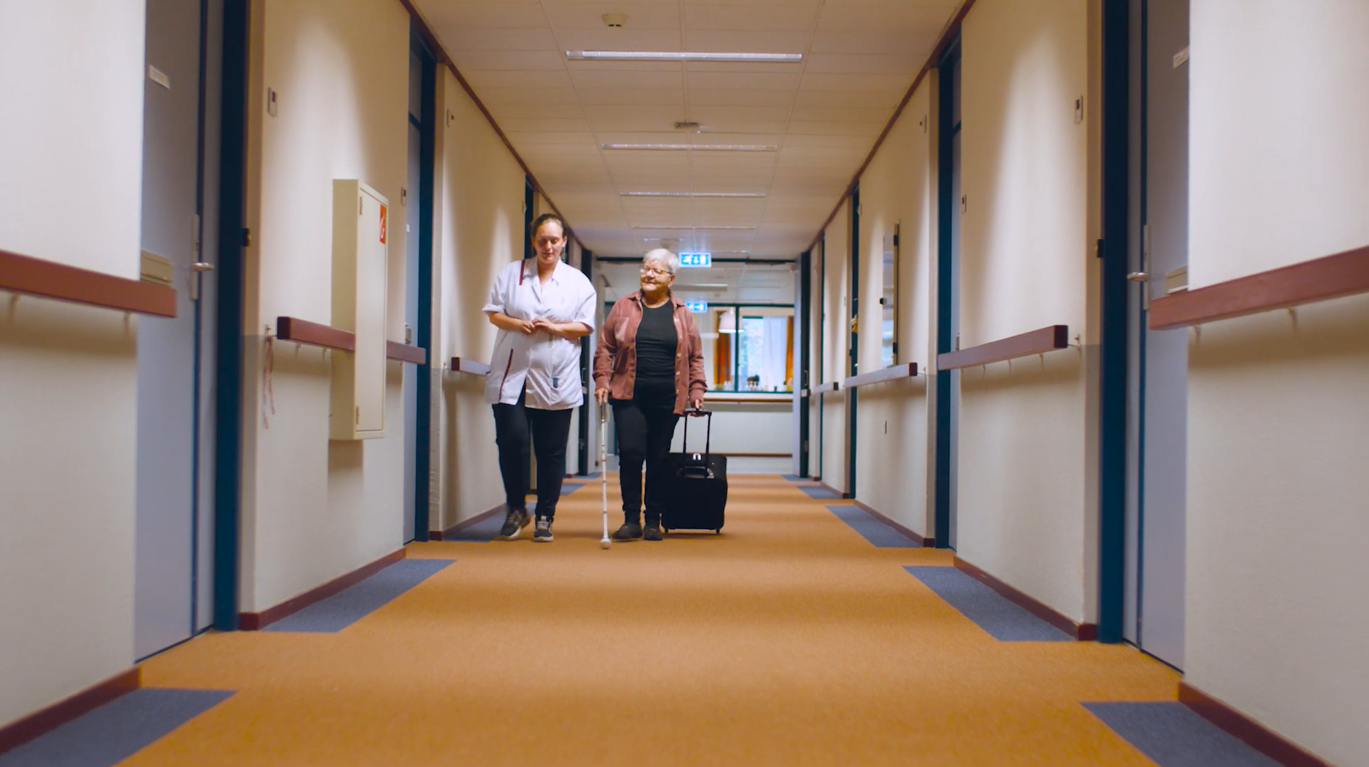 Cliënt met een taststok en een medewerker lopen samen door een hal van De Vlasborch Cliënt met een taststok en een medewerker lopen samen door een hal van De Vlasborch