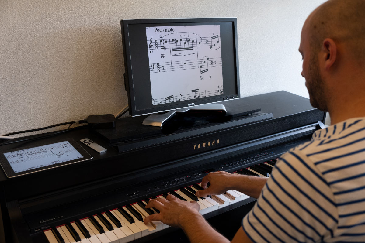 Cliënt van Visio Het Loo Erf speelt piano waarbij de bladmuziek vergroot is op een beeldscherm Cliënt van Visio Het Loo Erf speelt piano waarbij de bladmuziek vergroot is op een beeldscherm