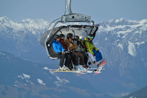 Zes mensen zitten in een skilift. Op de achtergrond de besneeuwde bergen.