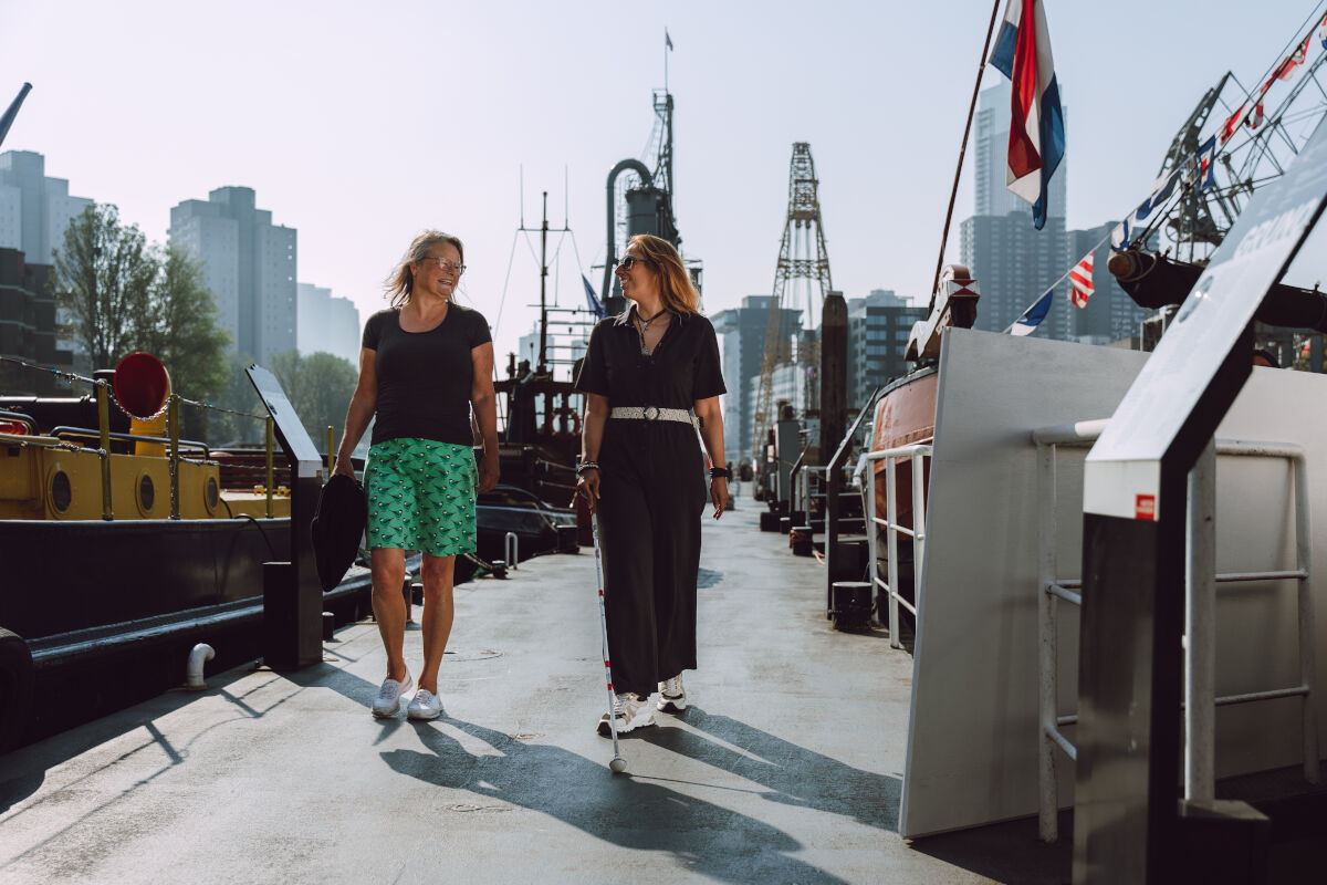Twee vrouwen lopen pratend op straat, waarvan een vrouw een taststok in haar handen heeft Twee vrouwen lopen pratend op straat, waarvan een vrouw een taststok in haar handen heeft
