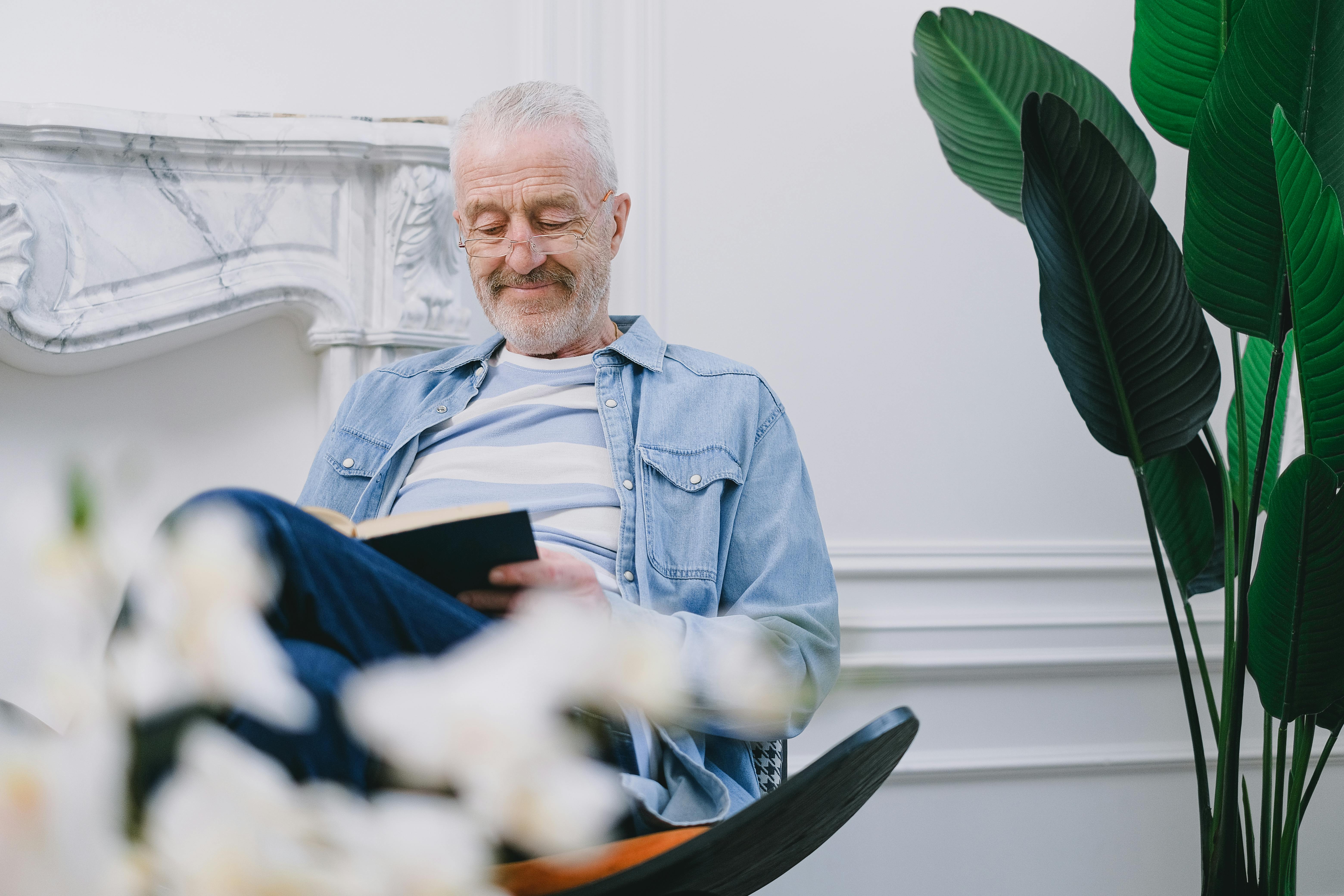 Man met grijs haar, bril, jeans en spijkeroverhemd leest een boek. Daarnaast zie je een marmeren schouw en een grote groene plant.