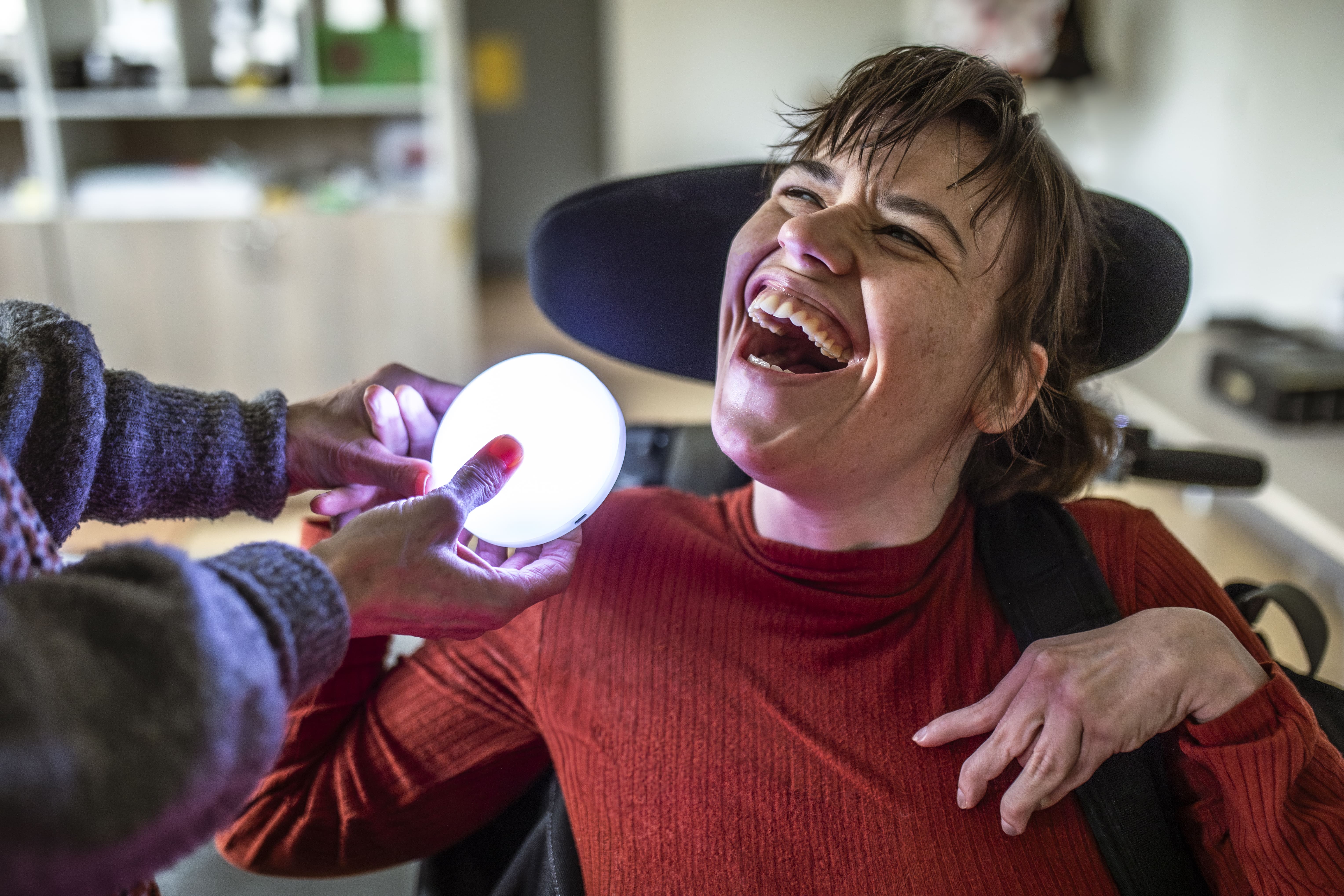 Een vrouw in een rolstoel heeft een grote glimlach op haar gezicht. Een begeleider houdt een lichtgevend object vast voor haar. Een vrouw in een rolstoel heeft een grote glimlach op haar gezicht. Een begeleider houdt een lichtgevend object vast voor haar.