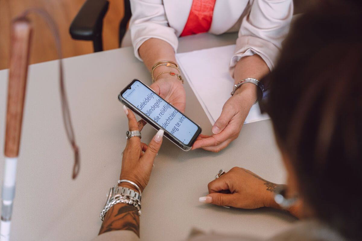 Twee paar handen houden een telefoon vast. Er staat vergrote tekst in beeld. Twee paar handen houden een telefoon vast. Er staat vergrote tekst in beeld.