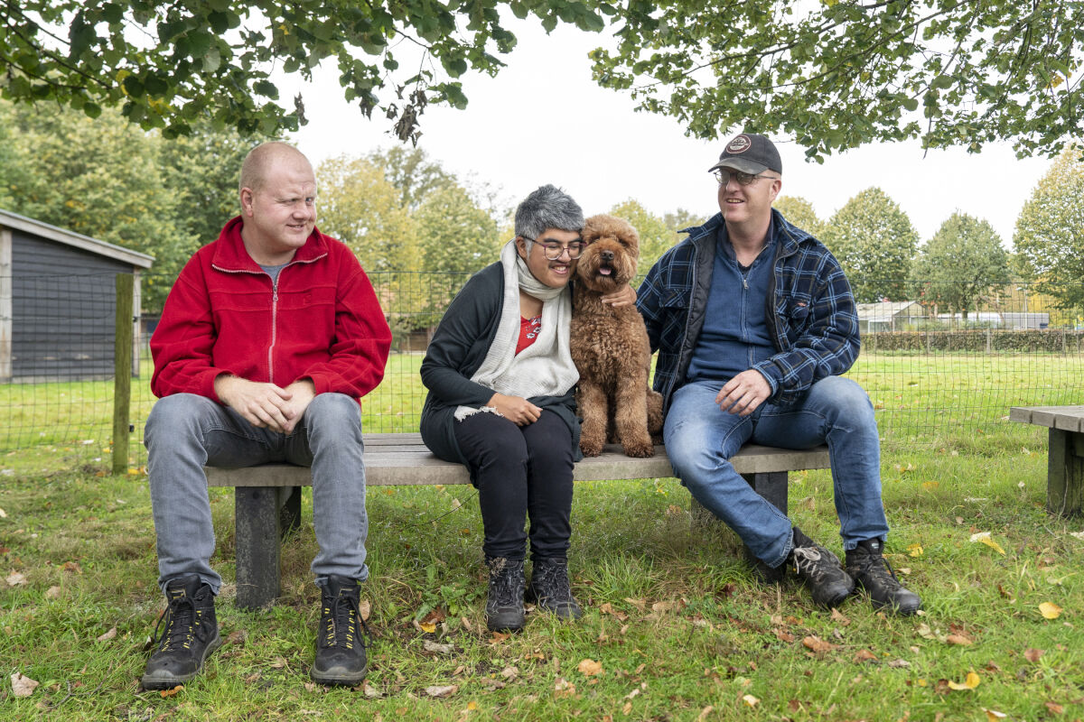 Drie cliënten van Visio zitten samen buiten op een houten bankje, één van hen knuffelt een hond Drie cliënten van Visio zitten samen buiten op een houten bankje, één van hen knuffelt een hond