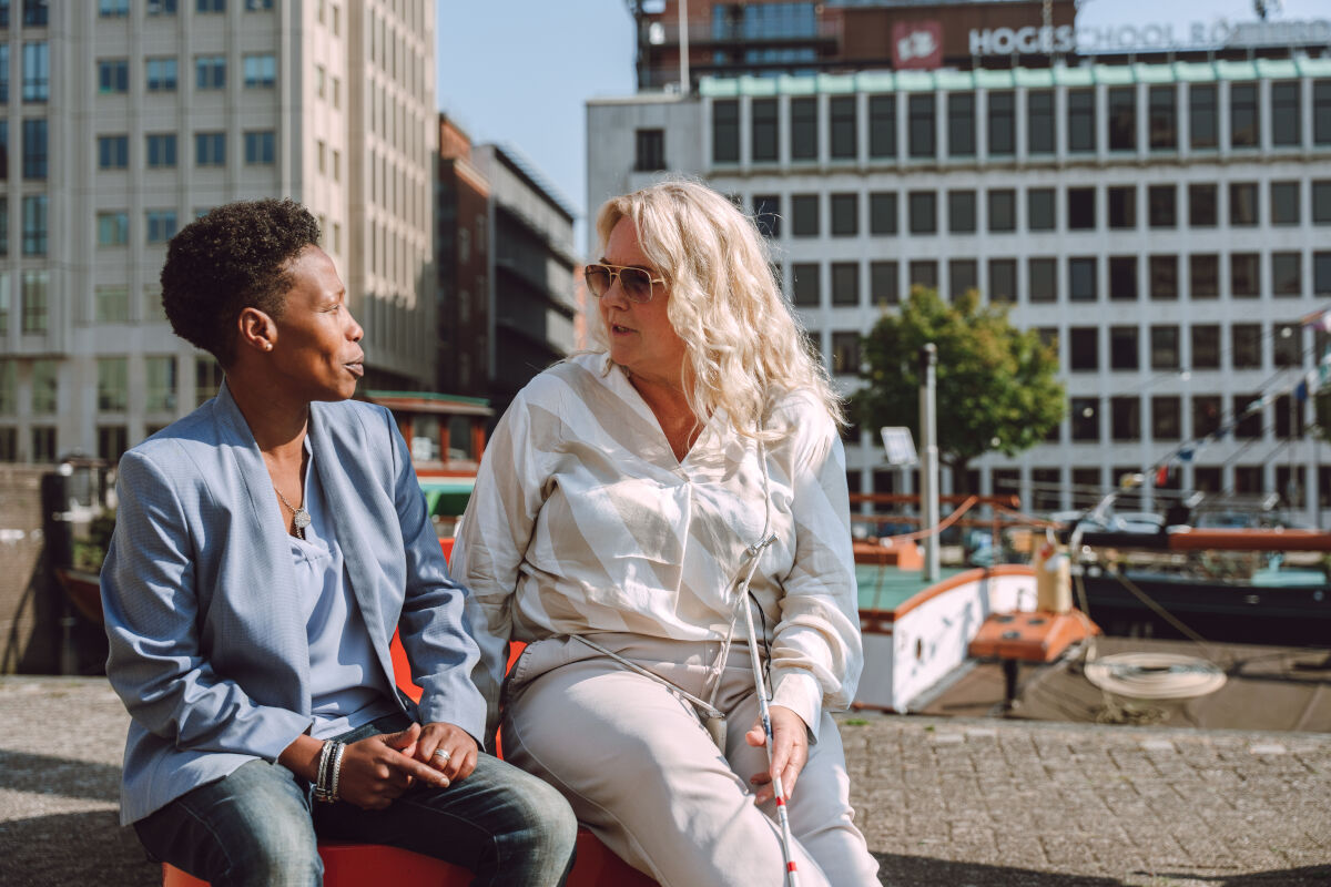 Twee dames zitten samen op een bankje en kijken elkaar aan. Één van de dames heeft een bril op en houdt een taststok vast.