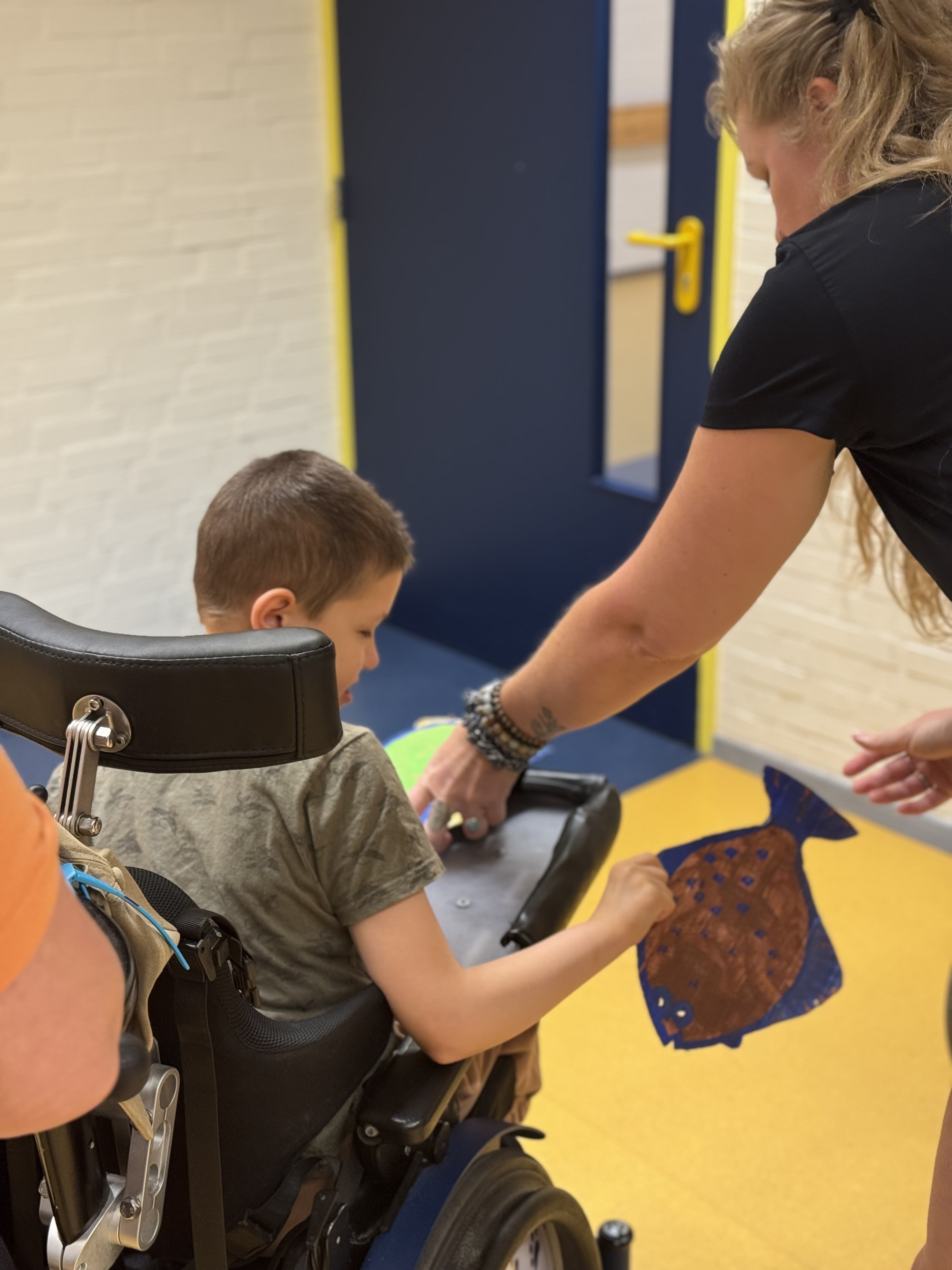 Begeleider helpt een kind in een rolstoel terwijl het kind een kleurrijke vis vasthoudt; in een gang met gele vloer en blauwe deur. Begeleider helpt een kind in een rolstoel terwijl het kind een kleurrijke vis vasthoudt; in een gang met gele vloer en blauwe deur.