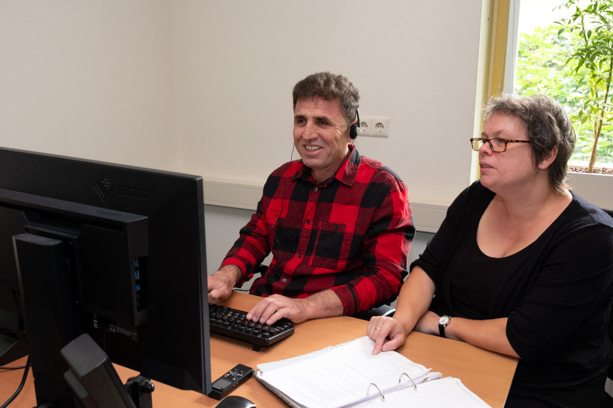 Cliënt en trainer van Visio Het Loo Erf zitten tijdens een ICT-training naast elkaar aan een bureau waarbij de cliënt oefent met het gebruik van de computer Cliënt en trainer van Visio Het Loo Erf zitten tijdens een ICT-training naast elkaar aan een bureau waarbij de cliënt oefent met het gebruik van de computer