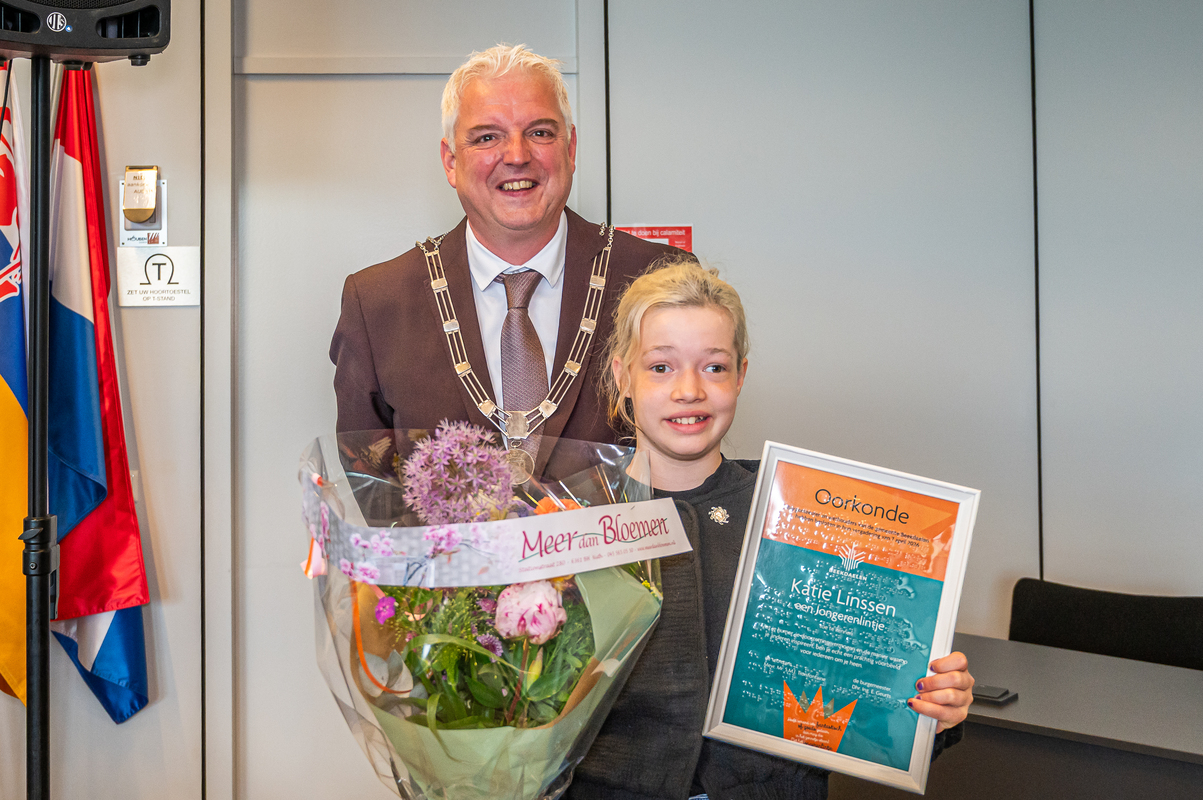 Burgemeester staat naast een kind dat een oorkonde en een grote bos bloemen vasthoudt in een binnenruimte. Burgemeester staat naast een kind dat een oorkonde en een grote bos bloemen vasthoudt in een binnenruimte.
